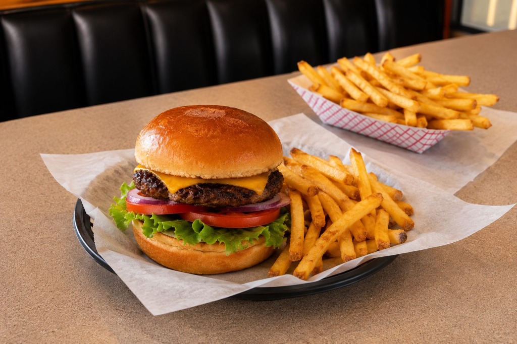 Big Spring BBQ - VLB Properties LLC - VLB Properties LLC Cheeseburger with lettuce, tomato, red onion and melted cheese on a glossy bun, with a side of golden fries on parchment-lined tray in a diner.",