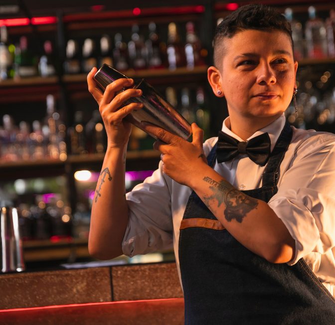 Bartender in a white shirt and bow tie shakes a cocktail shaker behind a bar, tattoos visible on forearms.