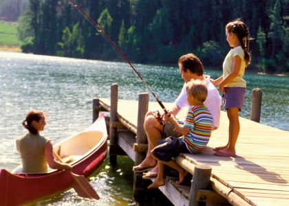 Family of four fishing from a wooden dock on a calm lake, with a canoe nearby and a forest backdrop.