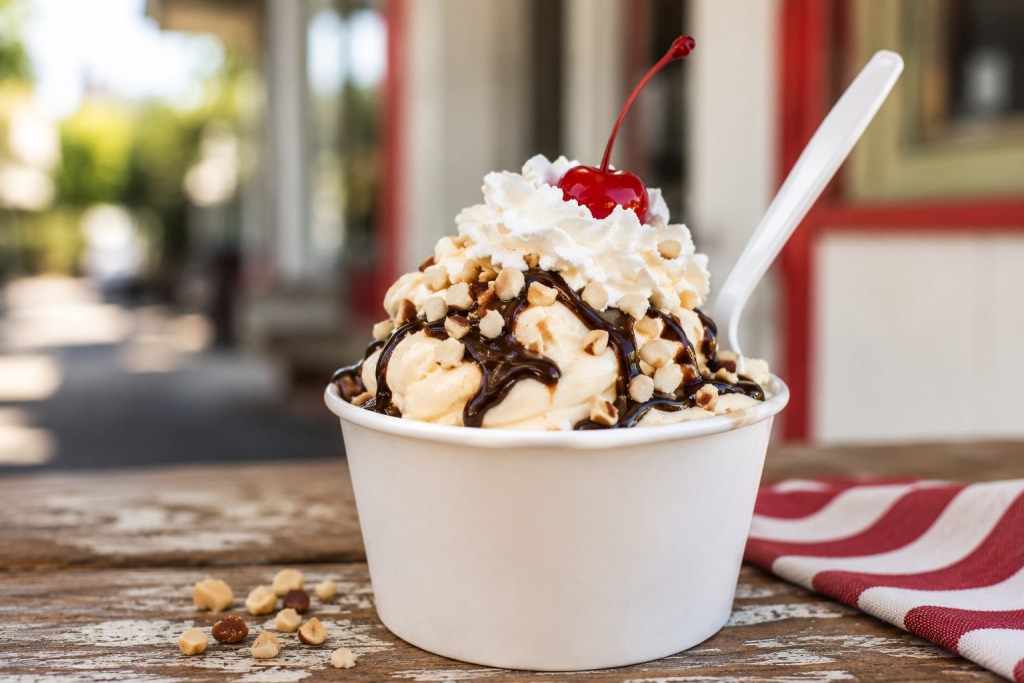 Pappys Original Frozen Custard - VLB Properties LLC - VLB Properties LLC Vanilla ice cream sundae in a white cup topped with whipped cream, chopped nuts, chocolate sauce, and a maraschino cherry, outdoors on a wooden table.