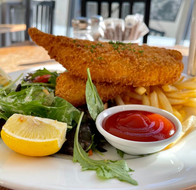 Plate of crispy fried fish fillet with French fries, greens, a lemon wedge, and a side of ketchup on a white plate.