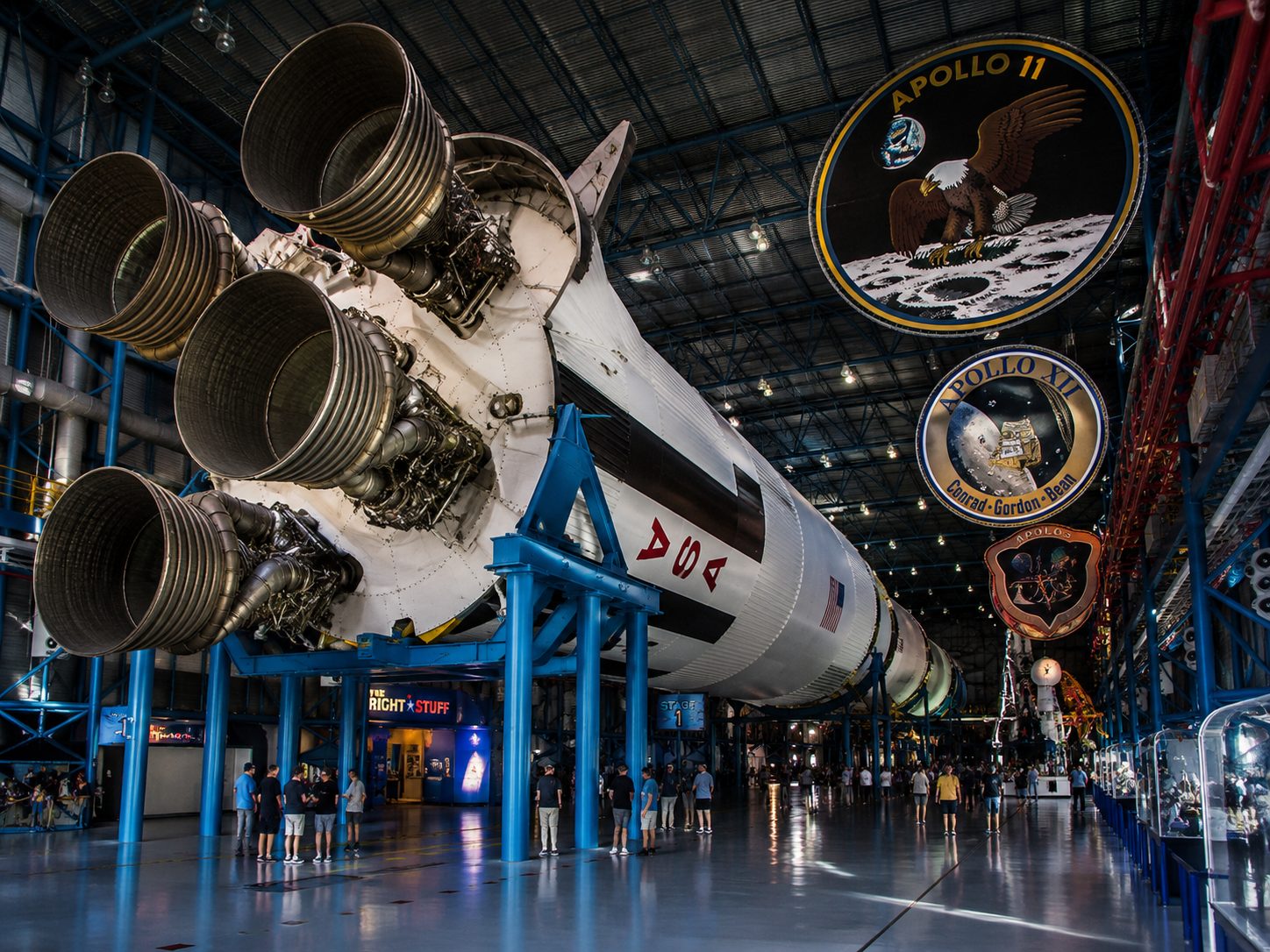 US Space Rocket Center - VLB Properties LLC - VLB Properties LLC Saturn V rocket on display in a museum hangar with visitors nearby and Apollo mission signs overhead and on the wall.