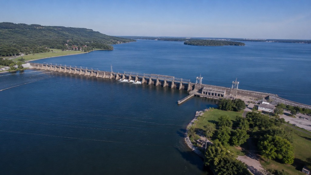 Wilson Lake + Wilson Dam - VLB Properties LLC - VLB Properties LLC Aerial view of a large concrete dam stretching across a blue lake, with forested hills on the left and a small island in the distance.